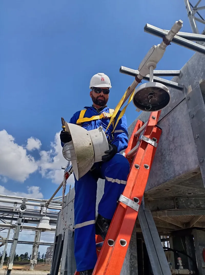 Técnico con braga azul, casco y arnés sobre una escalera naranja, sosteniendo una luminaria en una estructura metálica